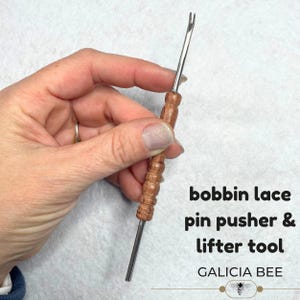 Puede incluir: Una herramienta para empujar y levantar alfileres de encaje de bolillos, con mango de madera y punta metálica. El texto "bobbin lace pin pusher & lifter tool" y "GALICIA BEE" son visibles.