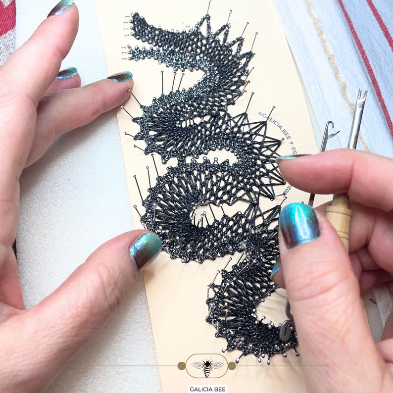 May include: A close-up of a black lace design pinned to a white board with a wooden tool and a needle. The lace design is in the shape of a dragon or a snake. The text "GALICIA BEE" is visible on the board.