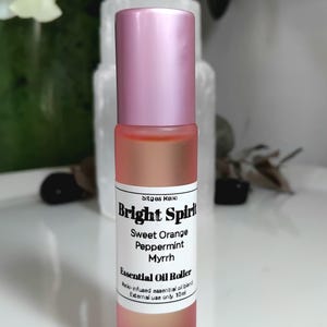 Könnte beinhalten: Eine 10ml ätherische Öl-Roll-on-Flasche mit einem mattierten rosa Glaskörper und einem metallisch rosa Verschluss. Das weiße Etikett trägt den Text Bright Spirit, Sweet Orange, Peppermint, Myrrh, Essential Oil Roller, Reiki-infused essential oil blend und External use only.