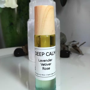Könnte beinhalten: Eine klare Glas-Roll-on-Flasche mit einem hellen Holzdeckel, die eine gelblich getönte Flüssigkeit enthält. Das weiße Etikett trägt den schwarzen Text DEEP CALM, Lavender, Vetiver, Rose und eine kleine Zeile mit use only Reiki-infused essential.
