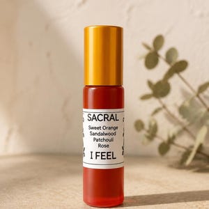 May include: A cylindrical orange glass roller bottle with a gold cap and a white label. The label features black text reading SACRAL, Sweet Orange, Sandalwood, Patchouli, Rose, and I FEEL, with small decorative black leaf icons on the sides.