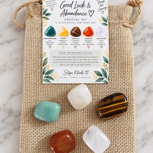 Op de afbeelding: Een jute buidel met trekkoord bevat vijf gepolijste edelstenen: groene aventurijn, gele citrien, bruin tijgeroog, oranje carneool en helder bergkristal. Een informatiekaart met de titel Good Luck & Abundance Crystal Set is inbegrepen.