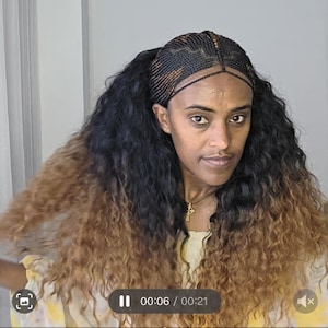May include: A woman with dark skin and long, wavy hair. The hair transitions from black to a light brown color. The top of the hair is styled in a braided pattern. She is wearing a gold necklace with a cross pendant.