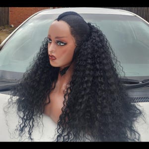 May include: A black curly human hair wig with a middle part and a high ponytail. The wig is styled in loose curls and is worn on a mannequin head.