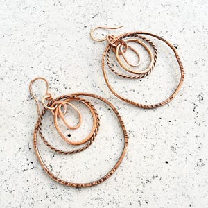 May include: Copper-colored earrings with a layered design. Each earring features multiple circular and oval shapes, with textured and smooth surfaces. The earrings have a hook closure and are displayed on a textured white surface.