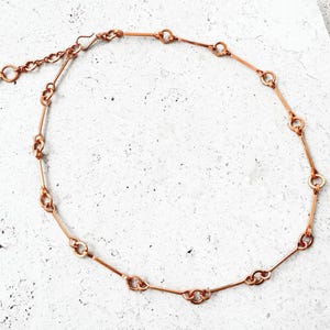 May include: A copper-colored necklace with a delicate chain design. The necklace features a series of interconnected links and a small, dark bead near the clasp. The necklace is displayed on a light-colored surface.
