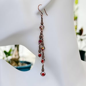 May include: A pair of long, dangling earrings with red and iridescent glass beads. The earrings are made with copper wire and have a simple, elegant design.
