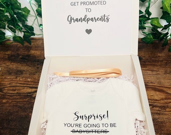 Puede incluir: Una caja de regalo blanca con una tapa blanca. La tapa tiene el texto "Only the best parents get promoted to Grandparents" con un pequeño corazón. La caja contiene un body blanco con el texto "Surprise! You're going to be babysitters... I mean Grandparents".