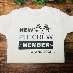 Peut inclure: Combinaison blanche pour bébé avec un texte noir qui dit "New Pit Crew Member Coming Soon..." avec un graphique de drapeau à damier noir et blanc.