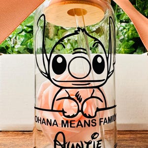 Puede incluir: Una botella de agua de vidrio transparente con tapa de bamb&uacute; y una pajita. La botella tiene una imagen de dibujos animados en blanco y negro de Stitch de Lilo & Stitch. El texto "Ohana means family" y "Auntie" est&aacute; impreso en la botella.