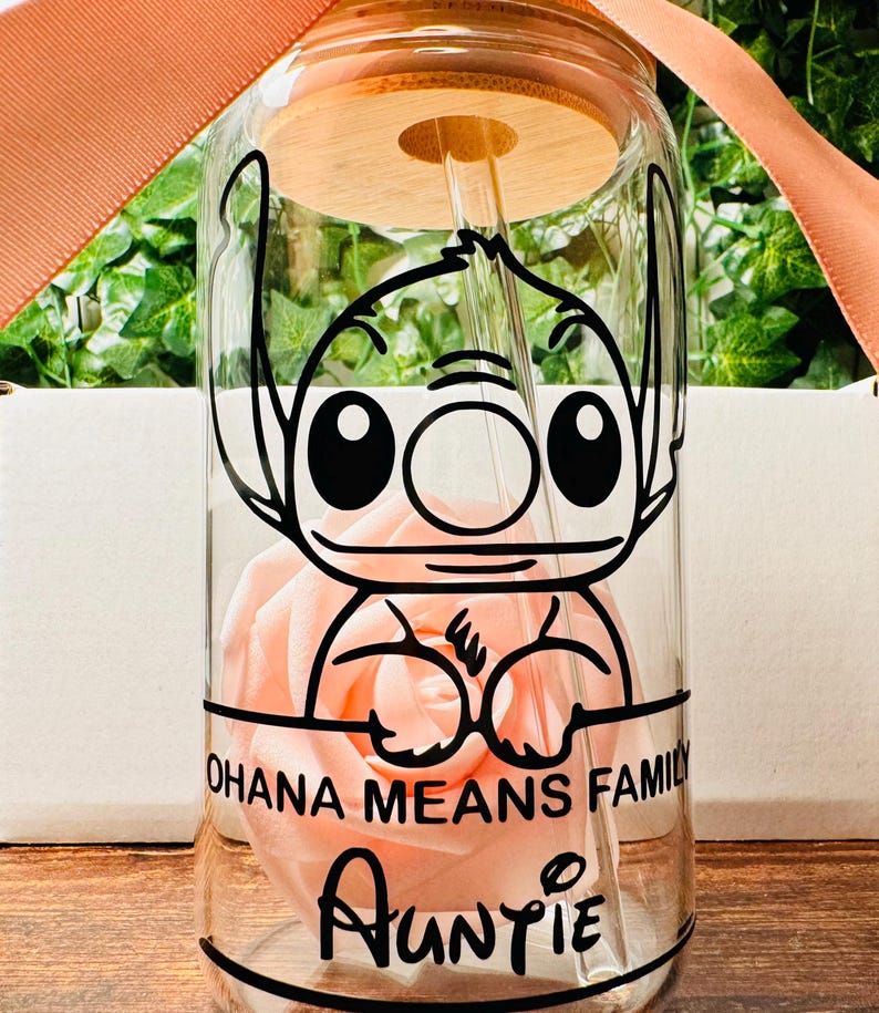 Puede incluir: Una botella de agua de vidrio transparente con tapa de bamb&uacute; y una pajita. La botella tiene una imagen de dibujos animados en blanco y negro de Stitch de Lilo & Stitch. El texto "Ohana means family" y "Auntie" est&aacute; impreso en la botella.