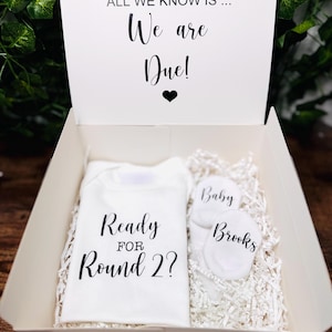 Op de afbeelding: Een witte geschenkdoos met een baby-aankondiging. Binnenin een witte romper met de tekst "Ready for Round 2?" en twee paar babysokjes. Op het deksel staat "It could be pink, it could be blue, all we know is... We are Due!"