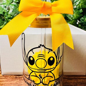 Puede incluir: Un vaso de vidrio transparente con un dise&ntilde;o de Stitch amarillo y negro. El vaso tiene una tapa de bamb&uacute; y una cinta amarilla atada alrededor. El texto "Ohana Means Family" y "Auntie" est&aacute; impreso en el vaso.