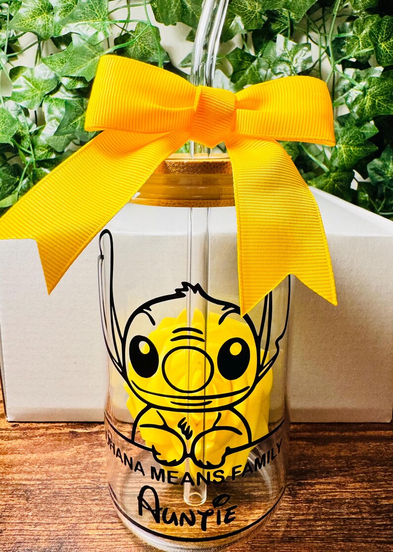 Puede incluir: Un vaso de vidrio transparente con un dise&ntilde;o de Stitch amarillo y negro. El vaso tiene una tapa de bamb&uacute; y una cinta amarilla atada alrededor. El texto "Ohana Means Family" y "Auntie" est&aacute; impreso en el vaso.