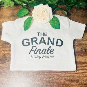 Puede incluir: Un body blanco para bebé con texto negro que dice "The Grand Finale" y "Julio 2025" con un corazón a cada lado de la fecha.
