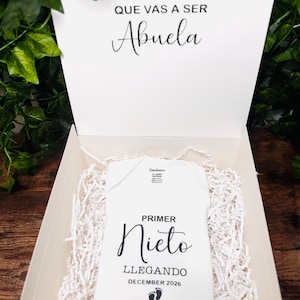 May include: A white gift box with a baby onesie inside. The onesie has the words "PRIMER Nieto LLEGANDO DECEMBER 2026" printed on it. The box lid reads "UN PAJARITO ME DIJO QUE VAS A SER Abuela". The box is filled with white paper shreds.