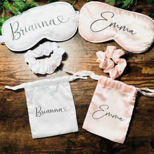 May include: Two sets of personalised sleep masks, scrunchies, and satin drawstring bags. One set is white with the name Brianna and a heart, and the other set is pink with the name Emma.