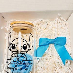 Puede incluir: Un tarro de vidrio con tapa de madera, que presenta un personaje de dibujos animados azul y una rosa azul. El tarro tiene el texto "We Ohana Coming Soon Auntie x 2 EST 2025" impreso en &eacute;l. El tarro est&aacute; empaquetado en una caja blanca con papel blanco triturado.