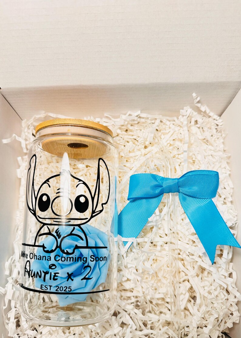 Puede incluir: Un tarro de vidrio con tapa de madera, que presenta un personaje de dibujos animados azul y una rosa azul. El tarro tiene el texto "We Ohana Coming Soon Auntie x 2 EST 2025" impreso en &eacute;l. El tarro est&aacute; empaquetado en una caja blanca con papel blanco triturado.