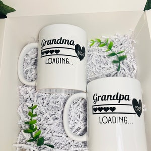 Op de afbeelding: Twee witte keramische mokken met zwarte tekst en harten. De mok links zegt "Grandma Loading... 2024" en de mok rechts zegt "Grandpa Loading... 2024".