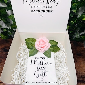 Könnte beinhalten: Eine weiße Geschenkbox mit einer rosa Rose darauf. Die Box ist geöffnet und im Inneren befindet sich ein weißes T-Shirt mit dem Text "I'm your Mother's Day Gift" und "See you in October 2025". Der Deckel der Box trägt den Text "This Mother's Day Gift is on Backorder".
