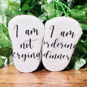 May include: A pair of white baby socks with black text. One sock reads "I am not crying" and the other sock reads "I am ordering dinner".