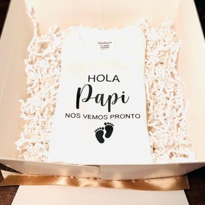 May include: White onesie with the text "Hola Papi Nos Vemos Pronto" and a pair of footprints in black.