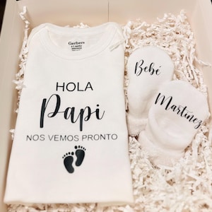 May include: A white onesie with the text "Hola Papi Nos Vemos Pronto" and two footprints.  The onesie is in a white gift box with white tissue paper.  There are two white baby mittens with the text "Bebé" and "Martinez" on them.