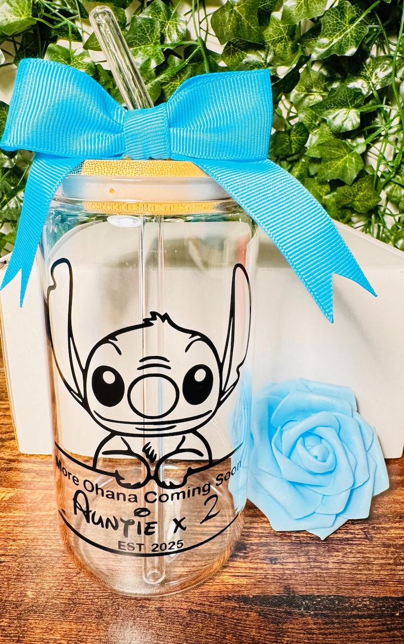 Puede incluir: Un vaso de vidrio transparente con una cinta azul atada alrededor de la parte superior. El vaso tiene una imagen en blanco y negro de Stitch de Lilo & Stitch. El texto en el vaso dice "More Ohana Coming Soon Auntie x 2 EST 2025".
