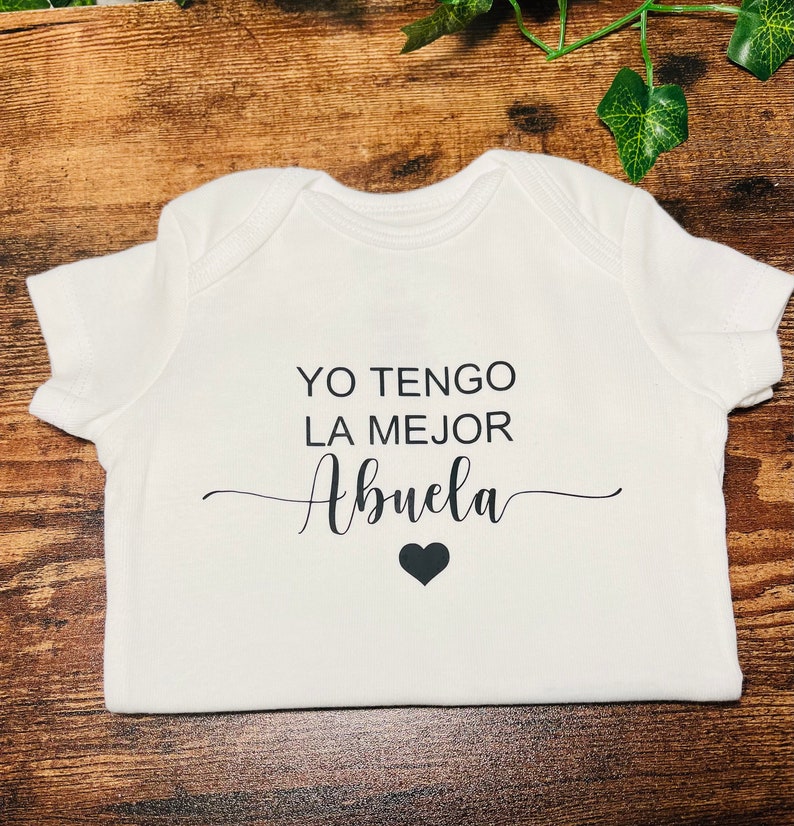 May include: White baby onesie with black text that reads "Yo tengo la mejor abuela" with a black heart below the text.