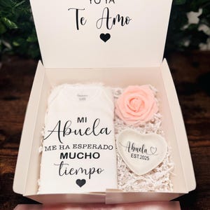 Peut inclure: Une boîte cadeau blanche contenant un body blanc qui dit "Mi Abuela Me Ha Esperado Mucho Tiempo" avec un cœur noir. La boîte contient également une rose rose et un plat blanc en forme de cœur qui dit "Abuela Est. 2025" avec un cœur noir.