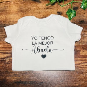 May include: White baby onesie with black text that reads "YO TENGO LA MEJOR ABUELA" with a small black heart below the text.