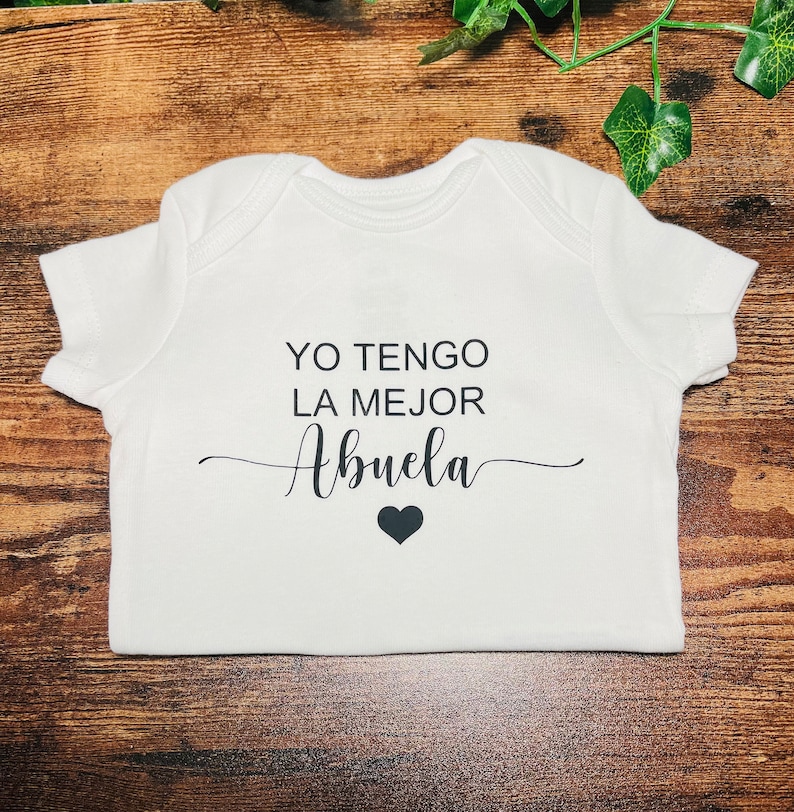 May include: White baby onesie with black text that reads "YO TENGO LA MEJOR ABUELA" with a small black heart below the text.