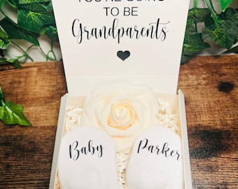 Puede incluir: Una caja blanca con una rosa blanca y dos calcetines de bebé blancos. La caja tiene una tarjeta en la parte superior que dice "¡Finalmente! Vas a ser abuelos". Un calcetín dice "Bebé" y el otro dice "Parker".