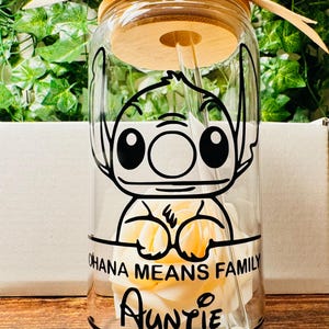 Puede incluir: Una botella de agua de vidrio transparente con tapa y pajita de bamb&uacute;. La botella presenta una imagen de dibujos animados en blanco y negro de Stitch de Lilo & Stitch sosteniendo una flor blanca. El texto "Ohana Means Family" y "Auntie" est&aacute; impreso en la botella.