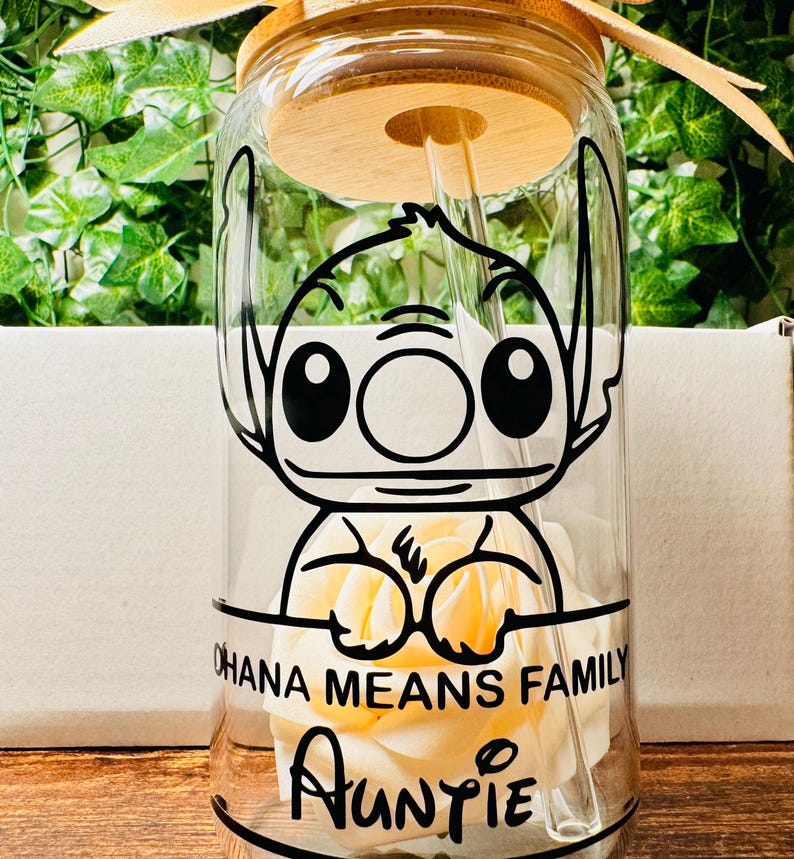 Puede incluir: Una botella de agua de vidrio transparente con tapa y pajita de bamb&uacute;. La botella presenta una imagen de dibujos animados en blanco y negro de Stitch de Lilo & Stitch sosteniendo una flor blanca. El texto "Ohana Means Family" y "Auntie" est&aacute; impreso en la botella.