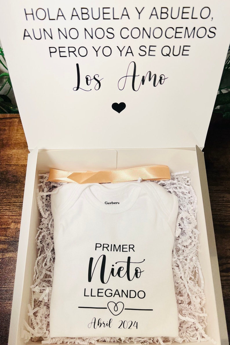 May include: A white onesie with the text "HOLA ABUELA Y ABUELO, AUN NO NOS CONOCEMOS PERO YO YA SE QUE Los Amo" and "PRIMER Nieto LLEGANDO Abril 2024" in black.