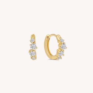 May include: A pair of gold hoop earrings with a cluster of clear, round-cut gemstones. The earrings have a polished finish and a simple, elegant design. The gemstones are arranged in a graduated pattern.