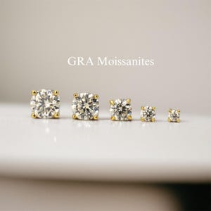 May include: Five gold stud earrings with clear, sparkling gemstones are displayed. The earrings vary in size, with the largest on the left and the smallest on the right. The text "GRA Moissanites" is above the earrings.
