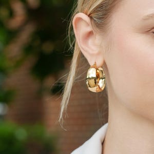 May include: Gold-colored, chunky hoop earrings. The earrings have a rounded, rectangular shape with a textured surface. The earrings are worn on the ear of a person with blonde hair.