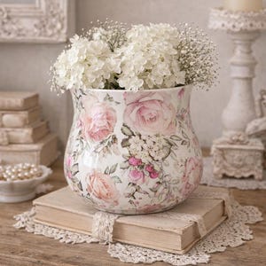 Peut inclure: Un vase décoratif rempli de fleurs blanches, orné d'un motif floral de roses roses et crème. Le vase est posé sur un livre vintage avec un napperon en dentelle, sur une surface en bois. Un bougeoir et d'autres objets vintage sont en arrière-plan.