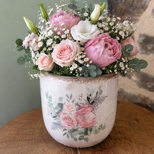 Peut inclure: Un arrangement floral dans un pot en céramique rond de couleur crème. Le bouquet présente des roses roses, des pivoines et des fleurs blanches, avec un feuillage vert. Le pot est décoré d'un motif floral. L'arrangement repose sur une surface en bois.