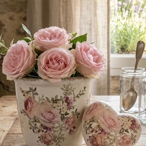 Peut inclure: Un arrangement floral avec des roses roses dans un pot en céramique décoratif. Un ornement en forme de cœur avec un motif floral assorti est posé sur une nappe en dentelle. La scène se déroule sur une table en bois, avec une fenêtre en arrière-plan.