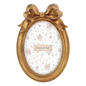 Peut inclure: Un cadre photo ovale doré surmonté d'un nœud décoratif. Le cadre contient un insert blanc avec un motif floral marron clair et le texte Clayre & Eef imprimé au centre sur une étiquette rectangulaire aux bords festonnés.
