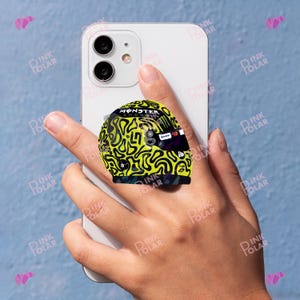May include: A phone grip featuring a miniature racing helmet design. The helmet is primarily yellow with black squiggly patterns and the "MONSTER ENERGY" logo. The grip is attached to a white smartphone, held by a hand against a blue background.