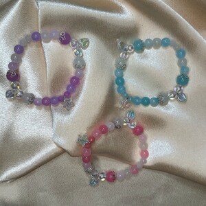 May include: Three bracelets made of colorful beads with silver charms. The bracelets are on a light pink fabric background.