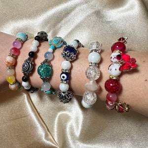 Op de afbeelding: Zes kleurrijke kralenarmbanden met verschillende bedels, waaronder een turquoise schildpad, een blauw boze oog en een rode bloem. De armbanden zijn gestapeld op de pols van een persoon.