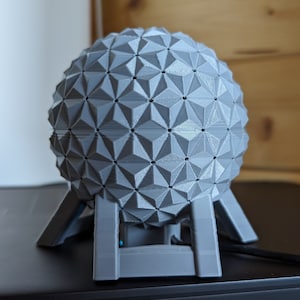 May include: A grey 3D printed model of the Spaceship Earth attraction at Epcot, featuring a geodesic sphere design with a base.