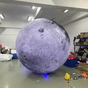 Op de afbeelding: Een grote opblaasbare bol, die op de maan lijkt, domineert de scène. De bol is een mix van paars en grijs, met gedetailleerde oppervlaktestructuren. De foto is binnenshuis genomen, met dozen en apparatuur op de achtergrond. De tekst "LAY-Z-DAYS EVENTS" is zichtbaar.