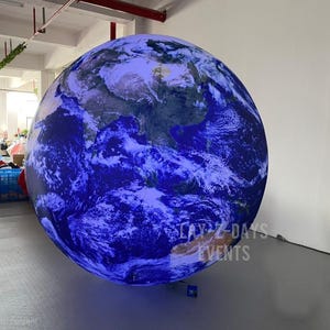 Op de afbeelding: Een grote opblaasbare wereldbol met een gedetailleerde weergave van de continenten en oceanen van de aarde. De bol is voornamelijk blauw en wit, met zichtbare landmassa's. De tekst "DAY 2-DAYS EVENTS" staat op de bol.
